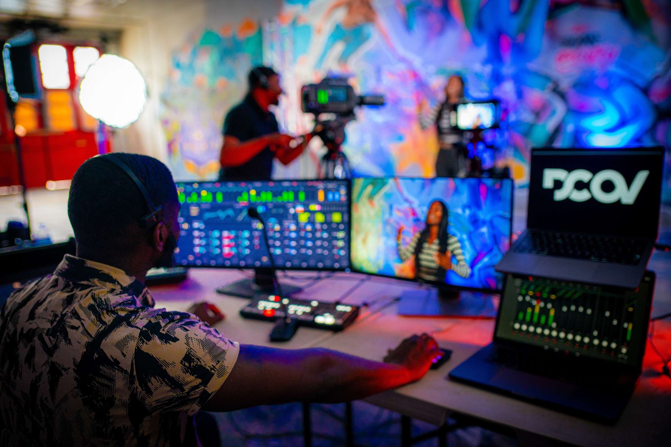 Video technician at a tri‑monitor control rig capturing a presenter against a colorful graffiti backdrop; softbox light, camera operator, and DCAV logo on a laptop visible